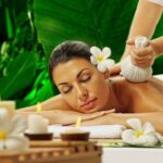 Woman receiving herbal compress therapy massage with plumeria flowers and candles against tropical green backdrop at Holistic Luxury Spa & Salon, Bhopal